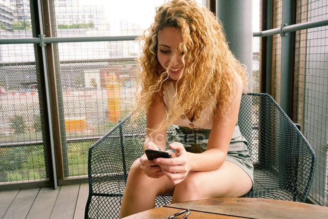Smiling curly woman chilling in modern waiting area sitting in metal chair and surfing mobile phone Texas — Stock Photo