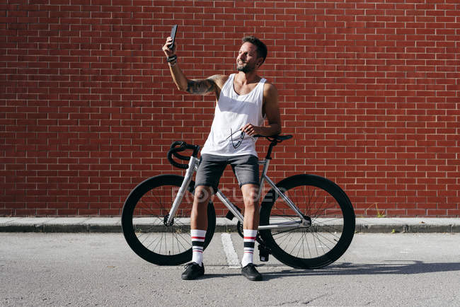 Bello ciclista in abbigliamento sportivo con smartphone mentre riposa sulla moto accanto al muro di mattoni rossi — Foto stock
