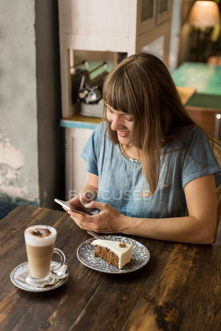 Loira alegre mensagem de digitação feminina no smartphone enquanto se senta à mesa com café e sobremesa no café acolhedor — Fotografia de Stock