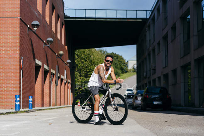 Sportive man in sunglasses wearing white sleeveless shirt and black shorts sitting on bicycle between buildings — Stock Photo