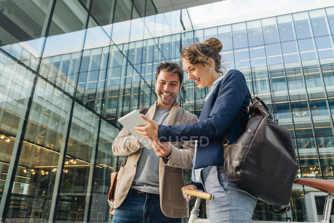 Gente de negocios optimista sonriendo y navegando en la tableta mientras está parado fuera del edificio moderno y descansando después del trabajo - foto de stock