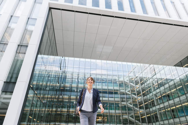 Baixo ângulo de mulher de negócios encantada sorrindo e andando fora do prédio de escritórios contemporâneo com parede de vidro — Fotografia de Stock