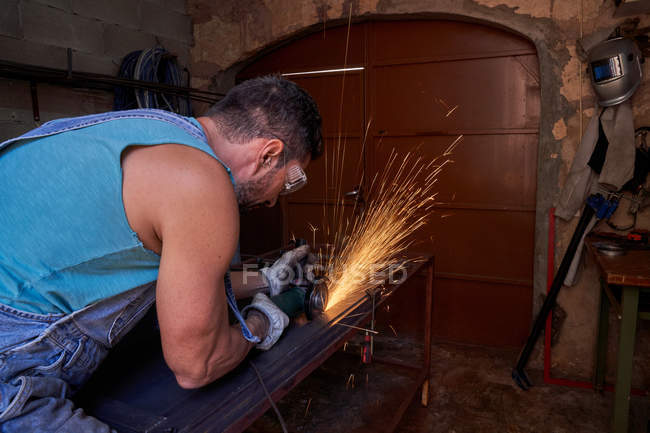 Seitenansicht des Arbeiters mit Schutzbrille und Handschuhen beim Schneiden von Metall mit einem Schleifer mit Funkenflug während der Arbeit in der Werkstatt — Stockfoto