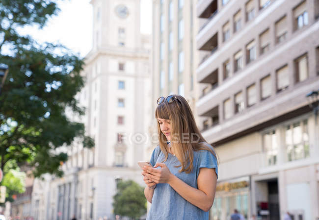 Bionda giovane donna allegra con occhiali da sole in piedi sulla strada della città e digitando il messaggio sullo smartphone — Foto stock