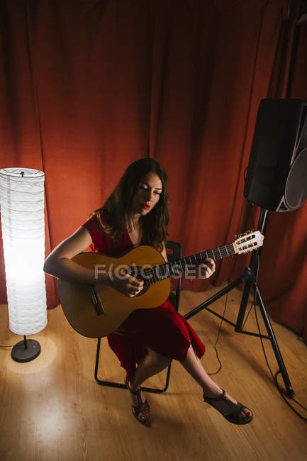 Charming artistic woman in red dress performing song playing on guitar in stage with warm light — Stock Photo