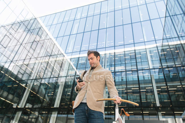 From below of pensive man making phone call while standing with bike against contemporary high rise business center — Stock Photo
