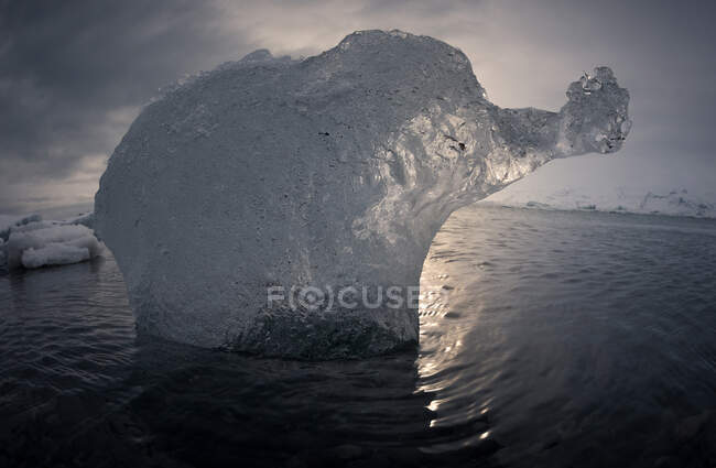Iceberg translúcido flutuando em água do mar frio contra céu noturno nublado na Islândia — Fotografia de Stock