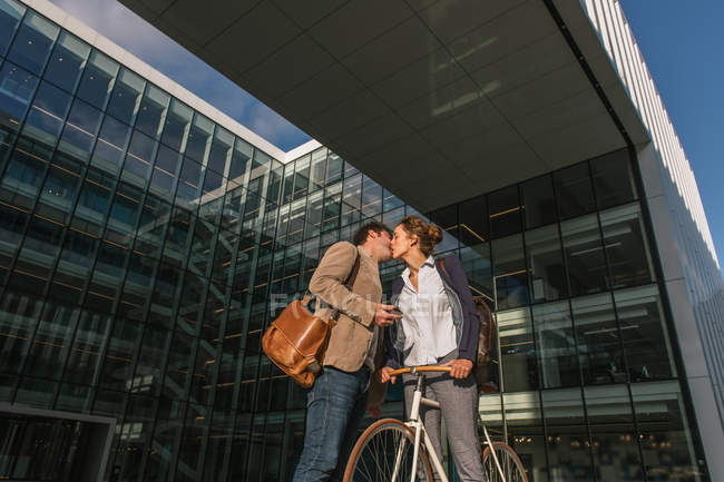 Homme et femme avec vélo s'embrassant tout en se tenant debout à l'extérieur de l'immeuble de bureaux sur la rue moderne de la ville — Photo de stock