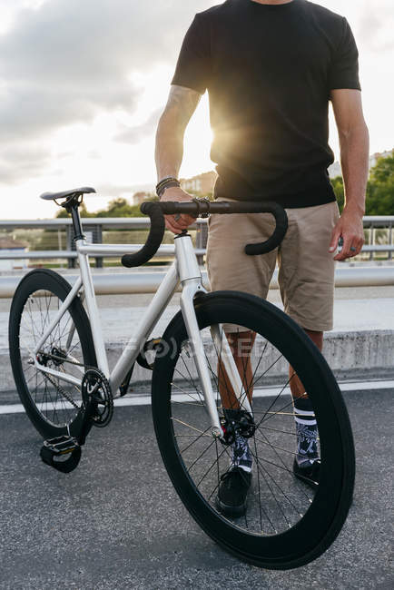 Imagem cortada do homem vestindo camisa preta e shorts bege de pé com bicicleta branca na ponte com edifícios da cidade no fundo — Fotografia de Stock