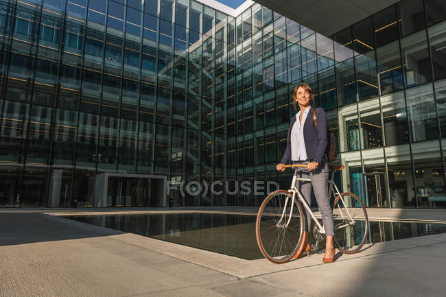 Вид сбоку восхищенной женщины-предпринимателя, улыбающейся и ездящей на велосипеде в солнечный день в центре современного города — стоковое фото