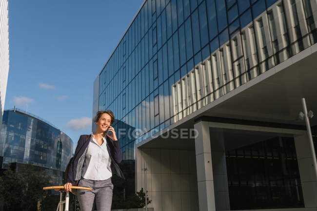 Mujer de negocios positiva con bicicleta sonriendo y hablando en el teléfono inteligente mientras camina fuera del edificio contemporáneo - foto de stock