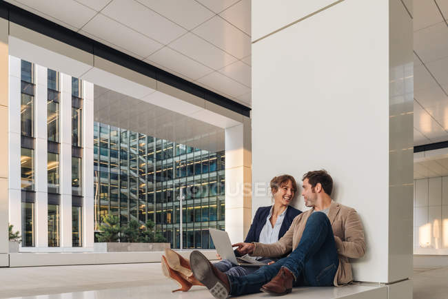 Delighted business people smiling and browsing laptop together while sitting outside modern building on city street — Stock Photo