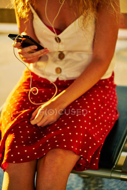 Imagen recortada de la mujer en auriculares escuchando música con teléfono móvil mientras se enfría en un banco de metal en el aeropuerto de Texas - foto de stock