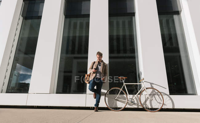 Serious entrepreneur with smartphone leaning on wall near bicycle on modern building and looking away in downtown — Stock Photo