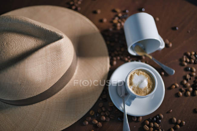 From above brown beverage with white foam in ceramic cup among coffee beans beside hat and empty mug on wooden table — Stock Photo