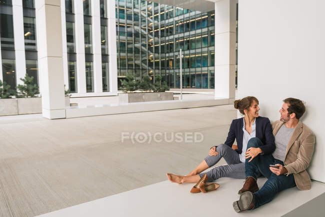 Веселая пара обнимает друг друга, просматривая смартфон, сидя возле современного здания — стоковое фото