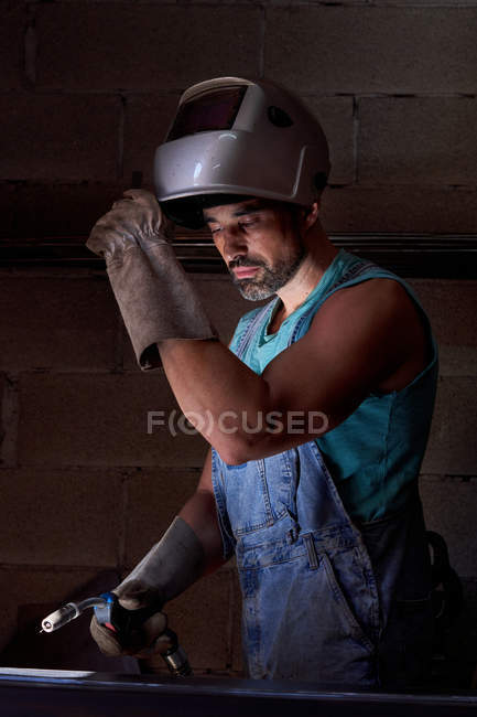 Profi-Mann mit Schweißmaske auf dem Kopf trägt Jeans-Overalls und Schutzhandschuhe am Arbeitsplatz und bereitet Instrument zum Löten vor, während er wegschaut — Stockfoto
