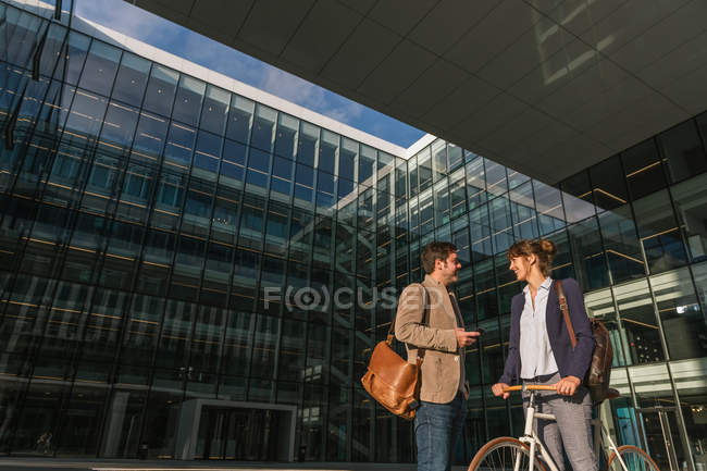 Alegre hombre y mujer con bicicleta sonriendo y mirándose el uno al otro fuera del edificio de oficinas en la calle moderna de la ciudad - foto de stock