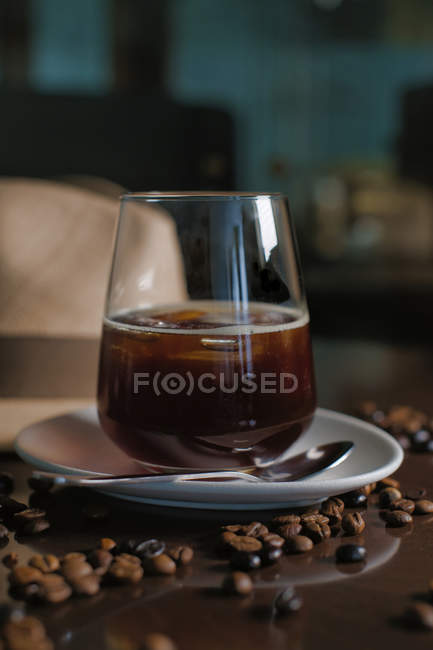 Deliciosa bebida marrón aromática en vidrio en platillo redondo entre granos de café asados al lado del sombrero en la mesa brillante - foto de stock
