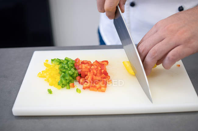 Manos de cocinero en uniforme moliendo pimientos rojos verdes amarillos con cuchillo grande en pizarra blanca - foto de stock