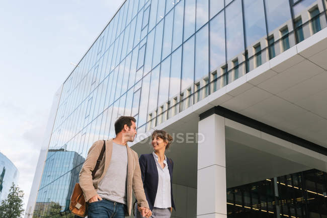 Знизу щаслива пара колег тримає руки посміхаючись і йдучи разом за межами сучасної будівлі — стокове фото