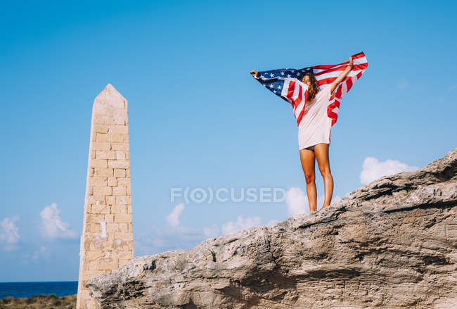 Fröhlich gebräunte Frau im Urlaub steht auf Klippen und hält amerikanische Flagge neben gerocktem Obelisk — Stockfoto