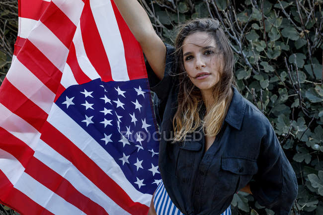 Alegre encaracolado fêmea hipster em casual camisa preta com bandeira americana olhando na câmera em arbustos verdes — Fotografia de Stock