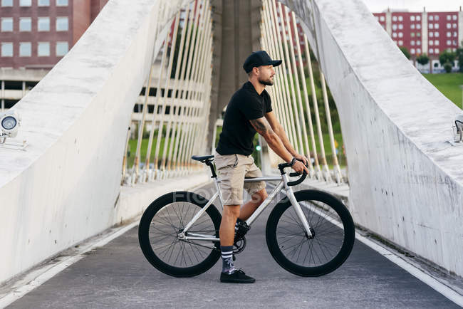 Hombre barbudo adulto feliz en gorra negra con camisa negra y pantalones cortos beige de pie con bicicleta cruzando pasarela en la ciudad mirando hacia otro lado - foto de stock