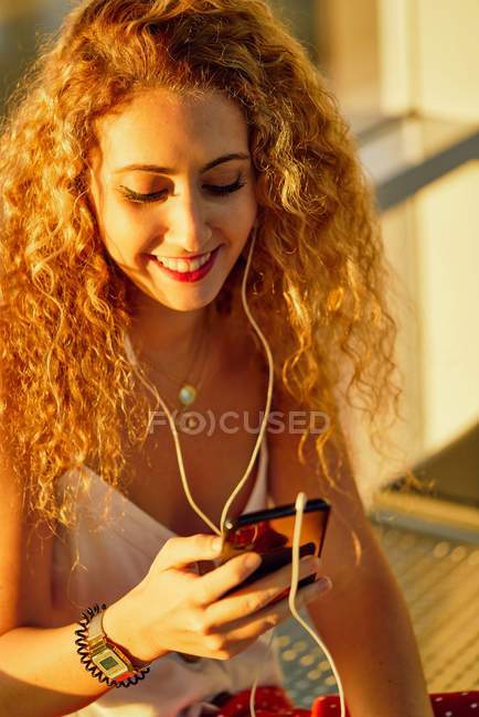 Pleasant cheerful woman in earphones listening to music with mobile phone while chilling on metal bench in airport of Texas — Stock Photo