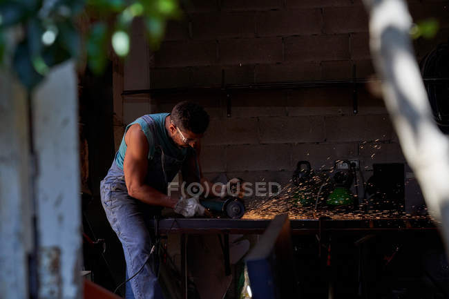 Ansichten von Baumzweigblättern eines Arbeiters in Schutzbrille und Handschuhen beim Schneiden von Metall mit einem Schleifer mit Funkenflug während der Arbeit in der Werkstatt — Stockfoto