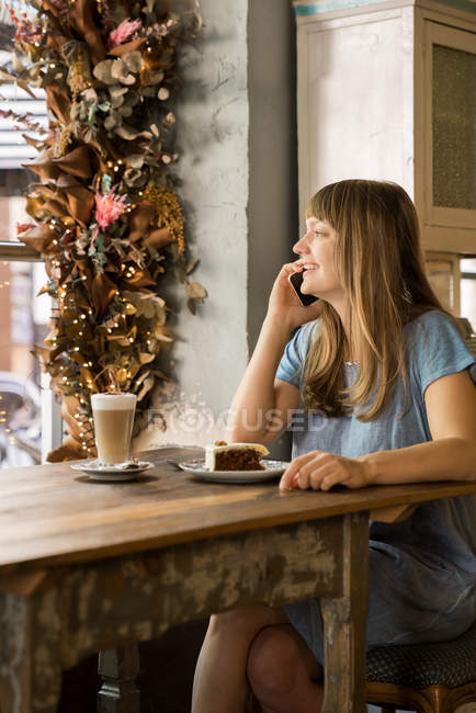 Bionda felice giovane femmina con frangia sorridente e parlando su smartphone in accogliente caffè — Foto stock