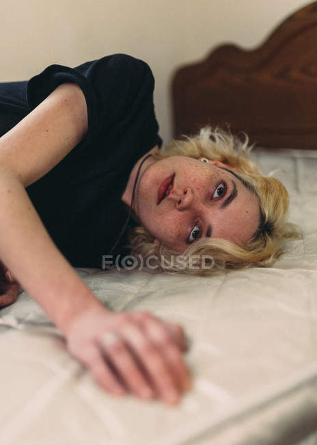 Pretty young blond freckled female with brown eyes in casual black shirt looking away and contemplating while resting in bed — Stock Photo