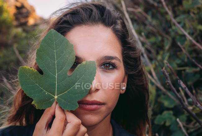 Peaceful young woman relaxing in nature while covering face with green leaf of fig and looking in camera — Stock Photo