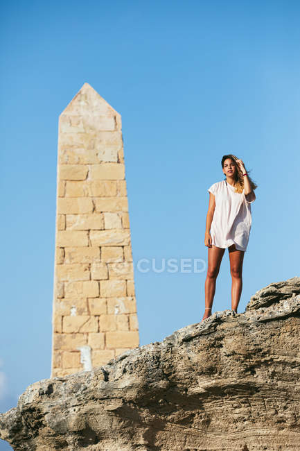 Кудрявая туристка в свободной легкой рубашке, смотрящая в камеру, стоя на скалах под скалистым обелиском — стоковое фото