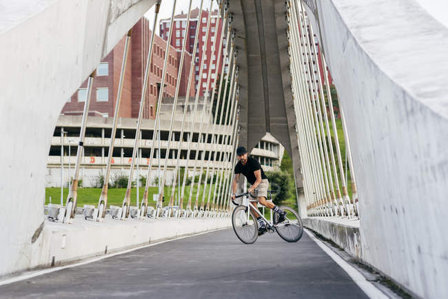 Adult bearded man in black cap wearing black shirt and beige shorts riding a bike across footbridge in city looking away — Stock Photo