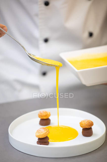 Mão de chef em tigela de retenção uniforme e derramando xarope amarelo de colher em prato branco com doces de chocolate — Fotografia de Stock