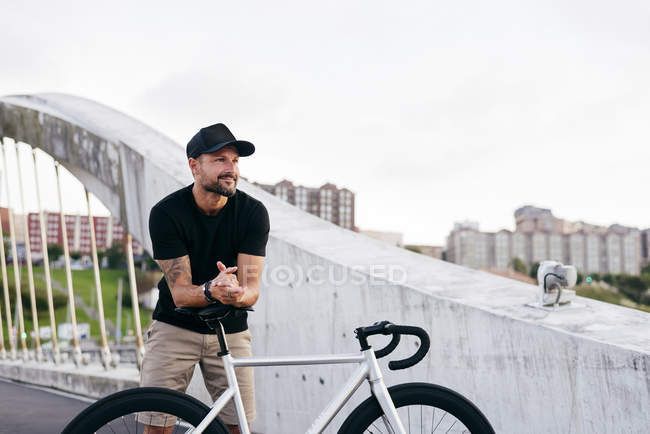 Hombre barbudo adulto feliz en gorra negra con camisa negra y pantalones cortos beige de pie apoyado en bicicleta sentarse a través de pasarela en la ciudad mirando hacia otro lado - foto de stock