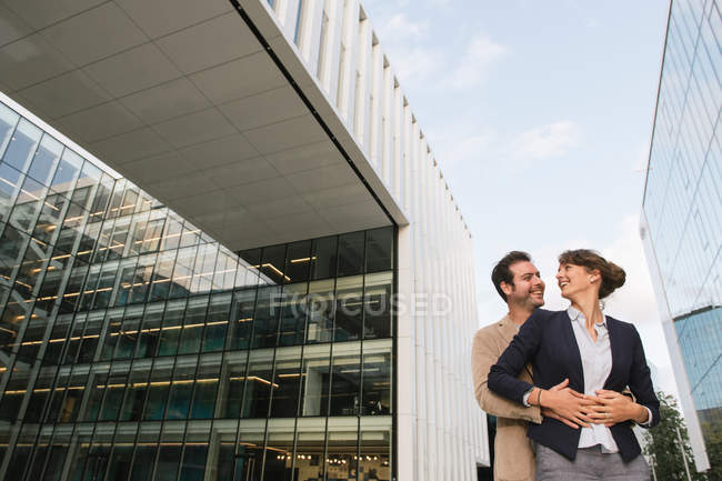 From below happy couple coworkers hugging while standing outside modern building on city street after work — Stock Photo