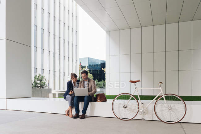 Delighted business people smiling and browsing laptop together while sitting outside modern building near bicycle on city street — Stock Photo
