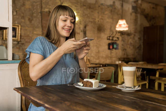 Loira alegre feliz fêmea com franja segurando smartphone sobre pedaço de bolo e sentado à mesa com café e sobremesa — Fotografia de Stock