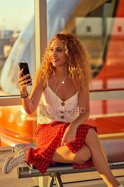 Приємна весела жінка в навушниках слухає музику за допомогою мобільного телефону і холоне на металевій лавці в аеропорту Техасу. — стокове фото