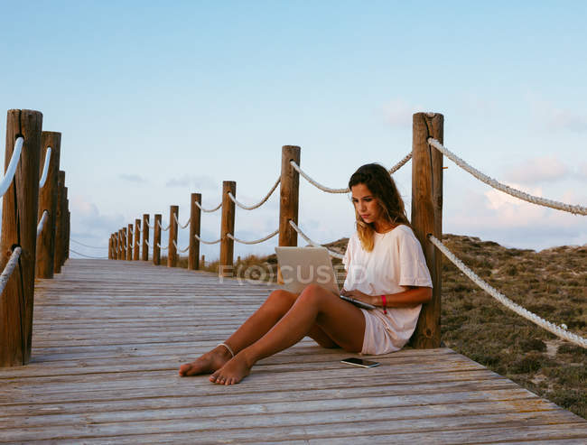 Concentrated female freelancer on vacation in white shirt sitting on empty bridge and working with laptop — Stock Photo