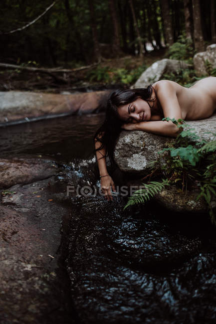 Mulher nua de cabelos longos pensive com olhos fechados deitado em pedra e água comovente em rio misterioso na floresta — Fotografia de Stock