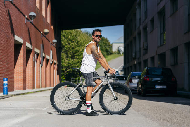 Sportive man in sunglasses wearing white sleeveless shirt and black shorts sitting on bicycle between buildings connected by bridge on summer sunny day — Stock Photo