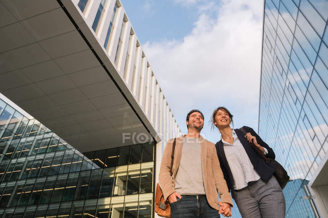 From below happy couple coworkers holding hands and walking together outside modern building on city street after work — Stock Photo