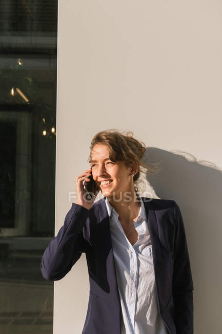 Optimistic businesswoman on phone in jacket smiling and looking away while leaning on building wall on city street — Stock Photo
