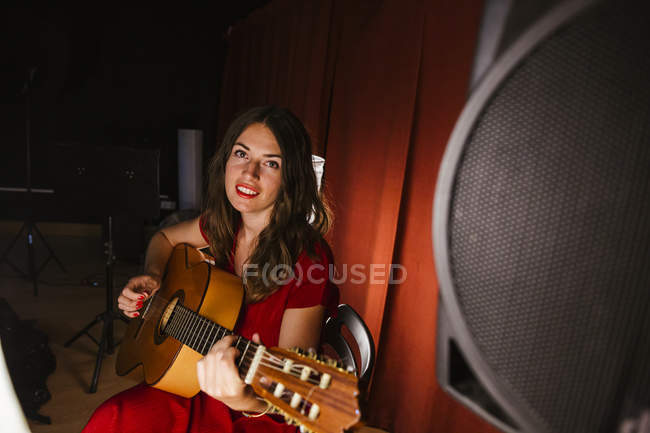 Очаровательная артистка в красном платье исполняет песню на гитаре на сцене с теплым светом — стоковое фото