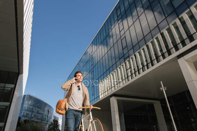 De baixo de homem pensativo que faz o telefonema ao estar com a bicicleta de encontro ao centro de negócios contemporâneo da elevação — Fotografia de Stock