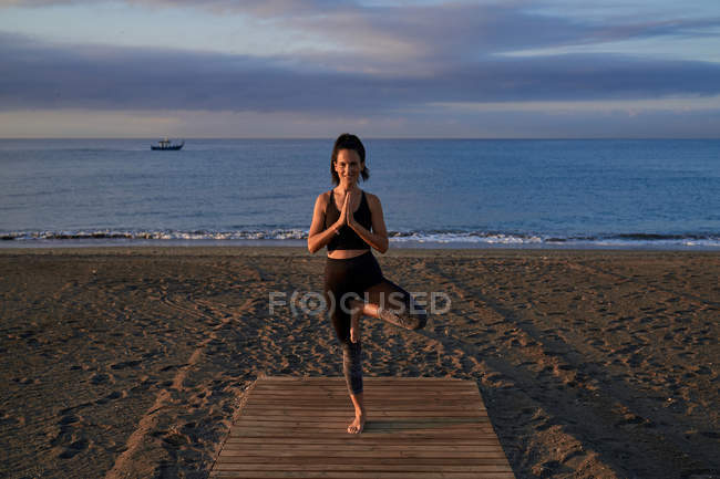 Happy slim female in sportswear standing in tree pose on sandy beach near waving sea in evening — Stock Photo