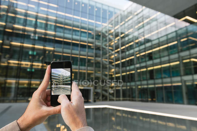 Мужские руки фотографируют со смартфоном прозрачный стеклянный фасад современного высотного бизнес-центра в центре города — стоковое фото